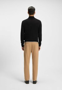 Maglione nero a maglia con colletto alto, abbinato a pantaloni beige con un leggero lucido e due tasche posteriori. Scarpe nere completano il look.