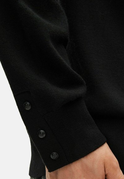 Manica di un maglione nero lavorato a maglia con tre piccoli bottoni neri vicino al polso, poggiata su una mano contro uno sfondo chiaro.