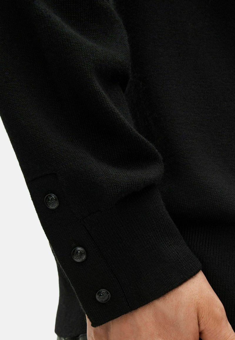 Schwarzer Strickpullover-Ärmels mit drei kleinen schwarzen Knöpfen nahe dem Handgelenk, der auf einer Hand vor einem hellen Hintergrund ruht.