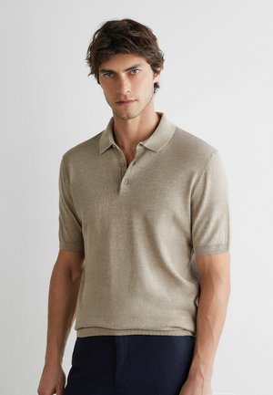 Junger Mann mit kurzen braunen Haaren, der ein beiges kurzärmeliges Poloshirt und dunkle Hose trägt, steht vor einem schlichten hellen Hintergrund.