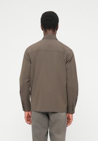 Καφέ πουκάμισο μακρυμ άνικο με ευθύ κόψιμο, μανσέτες με κουμπιά και επίπεδη γιακάς· λεία υφή υφάσματος και ελάχιστες λεπτομέρειες ραφής.