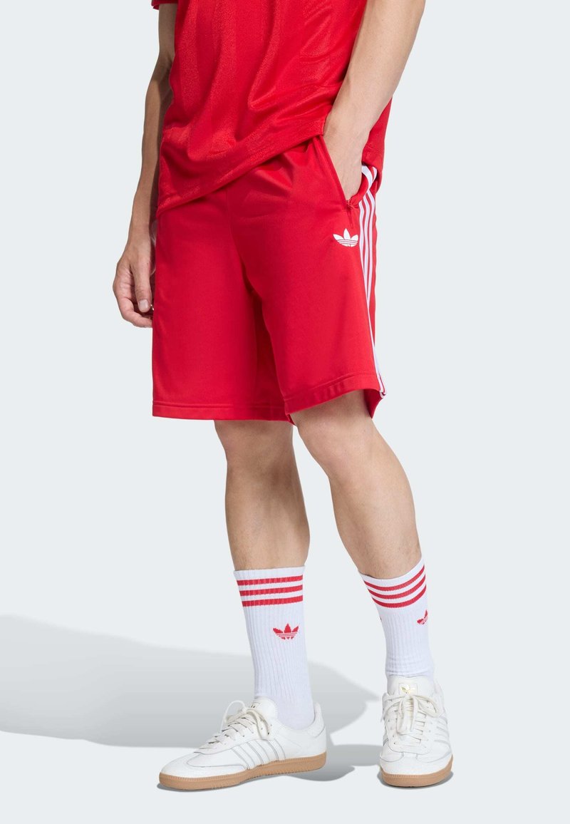 Shorts de sport rouges avec des bandes blanches sur les côtés, assortis à des chaussettes blanches avec des rayures rouges et un logo rouge. Portés avec des baskets blanches.