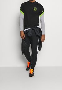 Schwarzes Nike-Sportshirt mit grünen Akzenten, grauen langen Ärmeln und Logo. Schwarze Hose mit weißen Streifen. Orangefarbene Stollenschuhe mit schwarzen Details.