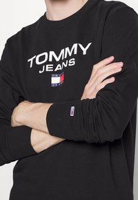 Sudadera negra de manga larga, hecha de algodón, con el texto en blanco "TOMMY JEANS" y un pequeño parche con el logo en la manga. Puños acanalados.