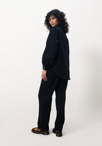 Chemise noire oversized et pantalon assorti en tissu doux, avec un ourlet légèrement incurvé et des poignets boutonnés. Des chaussures marron à motifs complètent le look.