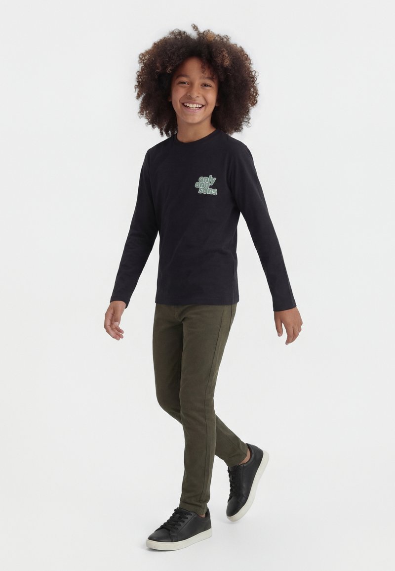 Otrok s kodrastimi lasmi, ki se smehlja, hodi, oblečen v črno majico z dolgimi rokavi z napisom, olivno zelene hlače in črne superge z belimi podplati.