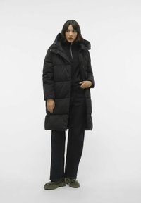 Manteau rembourré noir avec un design matelassé, doté d'une capuche amovible, d'une fermeture éclair et de poches latérales, associé à un jean foncé et des bottes vertes.