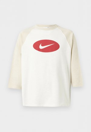 Jonge man met krullend haar die een wit en beige raglanmouwshirt draagt met een rood ovaal en witte Nike swoosh-logo.