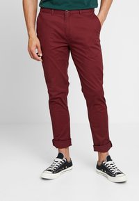 Mężczyzna w ciemnoczerwonych dopasowanych spodniach z podwiniętymi nogawkami i czarnych niskich trampkach z białymi sznurówkami, stojący z rękami w kieszeniach.
