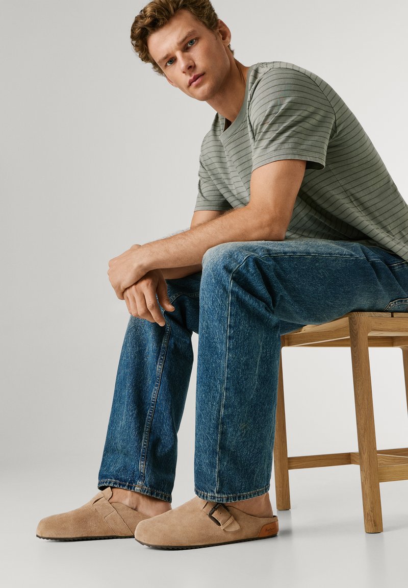 Νεαρός άνδρας που κάθεται σε ξύλινο σκαμπό, φορώντας γκρι ριγέ μπλούζα, μπλε τζιν και μπεζ παντόφλες με λεπτομέρεια αγκράφας.