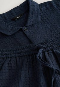 Blusa de color azul marino con textura que presenta un cuello con solapa, cierre de botones, cintura fruncida y detalle de cordón. Tejido de punto tipo gofre.