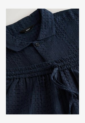 Granatowa bluzka o fakturowanej powierzchni, z kołnierzykiem, zapięciem na guziki, marszczonym pasem i szczegółem z troczkiem. Tkanina w splot wafelowy.