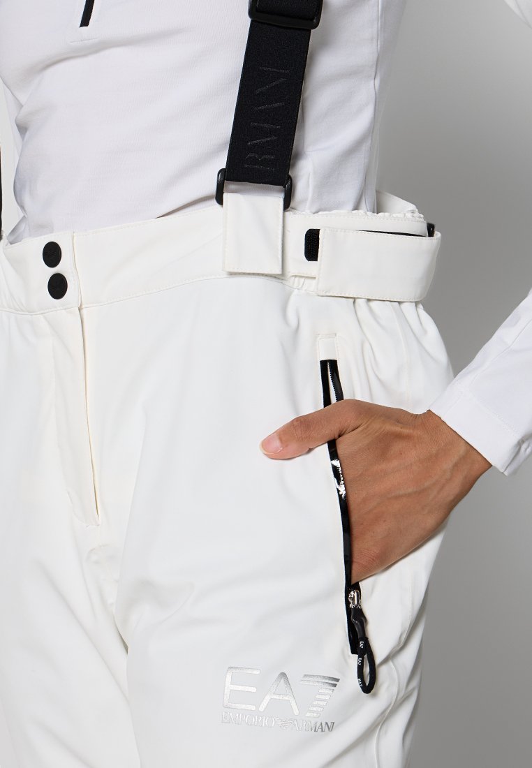 Pantaloni da sci impermeabili bianchi con bretelle regolabili nere, tasche laterali con zip e dettagli del logo argento sulla parte anteriore sinistra. Texture liscia.