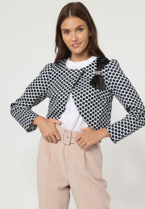Mujer con una chaqueta corta de lunares en blanco y negro con un lazo negro y pantalones beige de cintura alta ajustándose el cinturón.