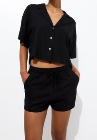 Camisa negra corta de botones con cuello, manga corta y tres botones blancos. Combinada con pantalones cortos negros de cintura elástica con bolsillos.