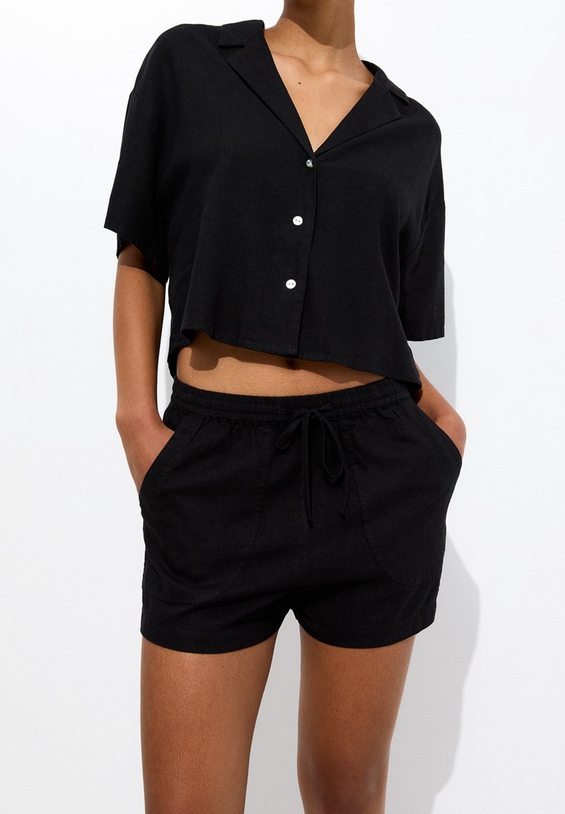 Camisa negra corta de botones con cuello, manga corta y tres botones blancos. Combinada con pantalones cortos negros de cintura elástica con bolsillos.