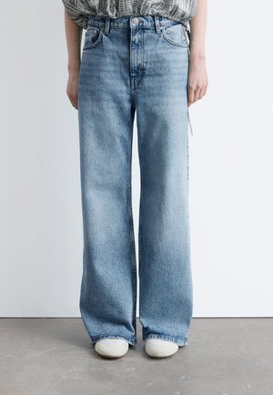 Personne portant un jean bleu clair taille haute à jambes larges et des chaussures blanches, debout contre un mur blanc uni sur un sol gris.