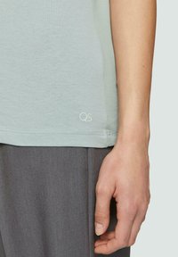 Hellgrünes T-Shirt mit kurzen Ärmeln und weicher Textur, mit einem kleinen "QS"-Logo nahe dem Saum. Getragen mit grauen Hosen.