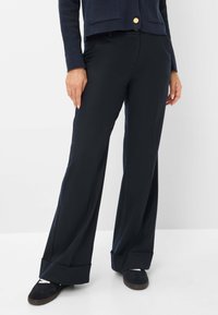 Femme portant un pantalon large bleu marine avec des revers, associé à des chaussures noires à enfiler, se tenant devant un fond blanc uni.
