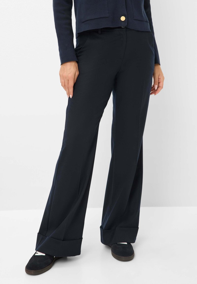 Femme portant un pantalon large bleu marine avec des revers, associé à des chaussures noires à enfiler, se tenant devant un fond blanc uni.