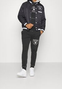 Černá saténová bomber bunda s bílými pruhy, na které je logo Raiders. Kombinována se šedými tepláky, které také zobrazují stejné logo, a bílými teniskami.