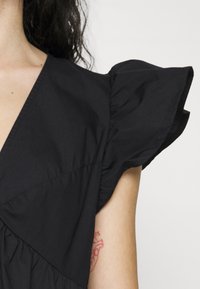 Gros plan d'une personne portant un haut noir à manches courtes à volants, montrant une partie de son cou, de son bras et un tatouage rouge à l'intérieur du bras.