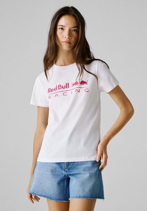 Young woman with long hair wearing white Red Bull Racing T-shirt and blue denim shorts against gray background.