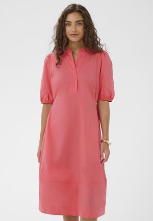 Hajlított hullámos hajú nő korallrózsaszín, térdig érő ruhát visel, amelynek könyökig érő puffos ujjai és részleges gombolórésze van, egyszínű háttér előtt áll.