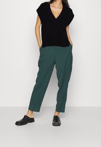 Schwarzer, gestrickter Pullover mit kurzen Ärmeln und V-Ausschnitt, kombiniert mit grünen, schmal geschnittenen Hosen und schwarzen Slippern.