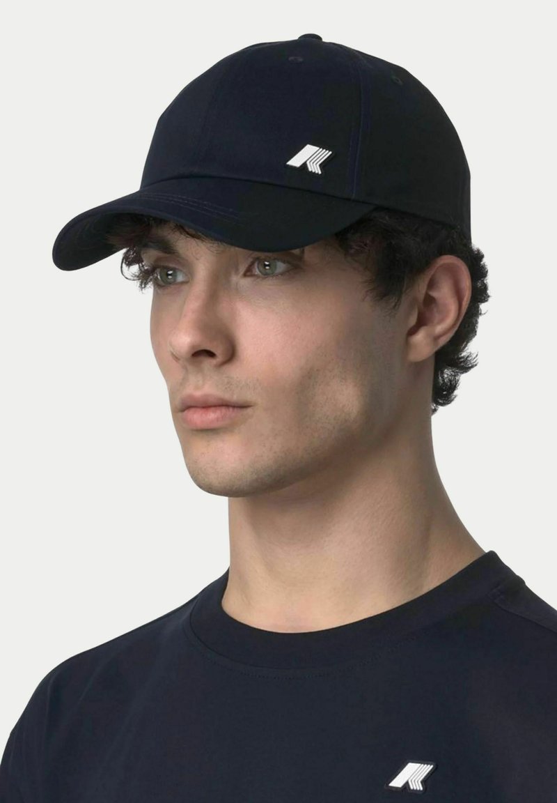 Jeune homme portant une casquette noire et une chemise noire, toutes deux ornées d'un petit logo géométrique blanc à l'avant.
