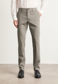 Mand iført beige lige pasform bukser, hvid skjorte med knapper og sorte loafers, står med den ene hånd i lommen på en neutral gulv.