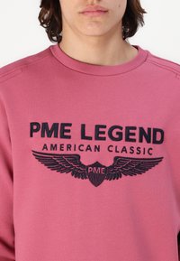 Różowa bluza z okrągłym dekoltem, z haftowanym czarnym logo z skrzydłami oraz napisem "PME LEGEND AMERICAN CLASSIC". Miękka tekstura.