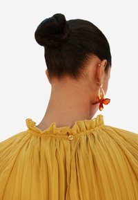 Blusa gialla plissettata con colletto arricciato, dotata di chiusura con bottoni sul retro; indossata con grandi orecchini floreali color ambra.