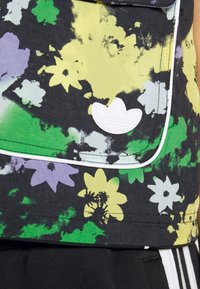 Ärmelloses Oberteil mit Blumenmuster, das gelbe, grüne und lila Blumen auf schwarzem Hintergrund zeigt, mit einem weißen Logodetail und kontrastierenden Paspeln.