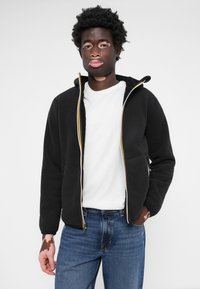 Svart fleece zip-up jakke med gule og hvite detaljer, brukt over en hvit T-skjorte, sammen med blå jeans. Jevn tekstur og casual passform.