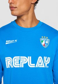 Mand iført en blå Replay FC sportshem med hvid Replay-logo og holdemblem, der viser tre gule stjerner, et italiensk flag og en blomst.