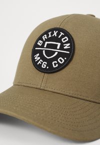 Șapcă de baseball verde măsliniu din țesătură texturată, cu un patch rotund negru pe care este imprimat în alb textul "BRIXTON MFG. CO." și un design simplu al logo-ului.