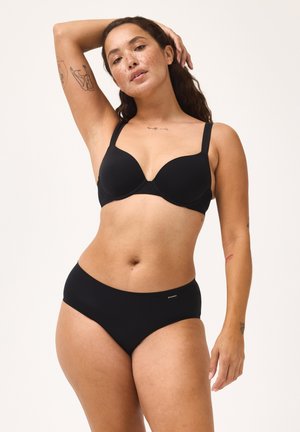 Donna con lentiggini e tatuaggi che posa indossando un reggiseno e slip neri, con un braccio sollevato, davanti a uno sfondo semplice.