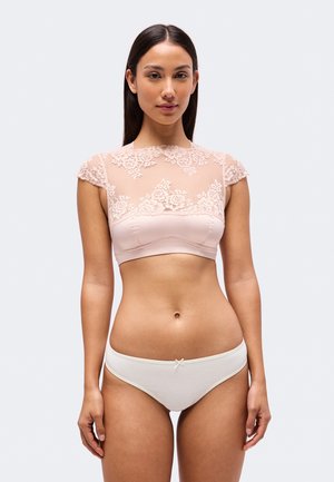 Une femme porte un crop top en dentelle rose pâle et une culotte blanche sur un fond clair et uni.