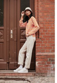 Kvinna som bär en beige bucket hat, långärmad tröja i orange och vit rand, beige byxor och vita sneakers, stående mot en tegelvägg.