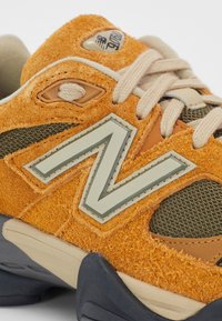Nærbillede af en orange og beige New Balance-sneaker med ruskindsstruktur, beige snørebånd og en stor "N"-logo på siden.