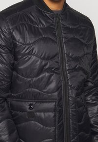 Giacca bomber imbottita nera con texture trapuntata ondulata, chiusura frontale con zip, tasca con bottone a scatto e colletto a coste.