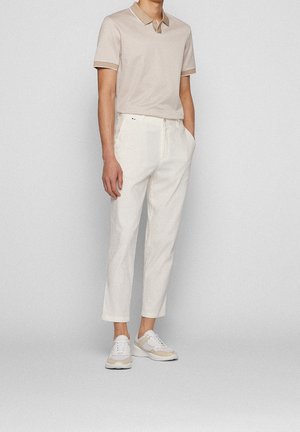 Mand iført beige kortærmet polo, hvide korte bukser og hvide sneakers, stående med hænderne i lommerne.