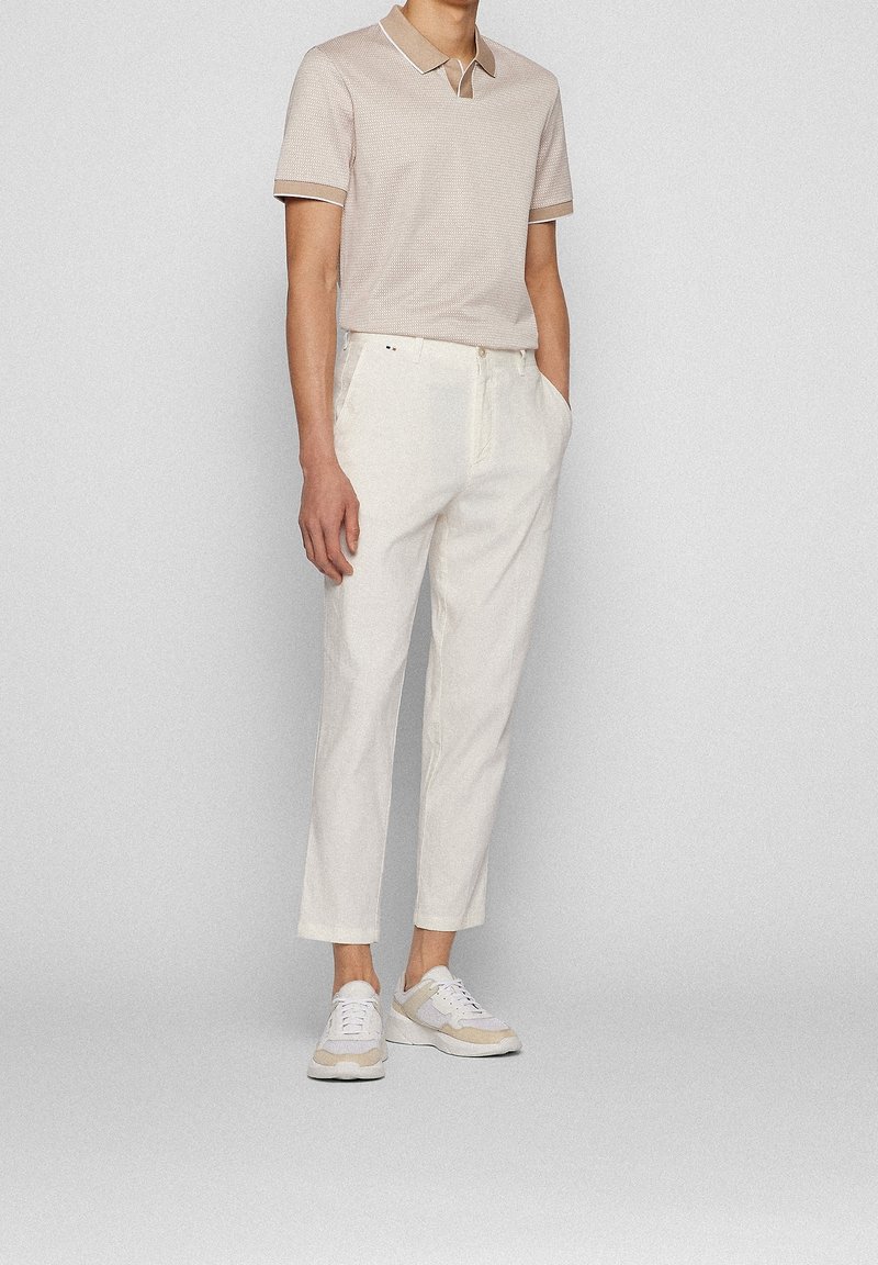 Homme portant un polo beige à manches courtes, un pantalon blanc court et des baskets blanches, debout les mains dans les poches.