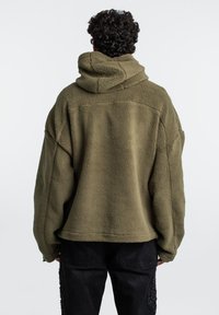 Sudadera de forro polar verde con un ajuste relajado, superficie texturizada, gran capucha y hombros caídos, combinada con pantalones de mezclilla negros con desgastado.