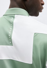 Άνδρας που φοράει πουκάμισο με μπλοκ χρωμάτων σε πράσινο και λευκό, με κατακόρυφο κείμενο "J.LINDEBERG" στην πλάτη κοντά στον γιακά.