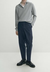 Jasnoszara koszulka polo z długim rękawem i małym kołnierzykiem, zestawiona z ciemnoniebieskimi zwężanymi spodniami i czarnymi sznurowanymi butami.