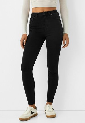 Personne portant un jean noir taille haute ajusté, un top blanc court à manches longues et des baskets blanches avec logos Swoosh noirs, debout sur un sol blanc.