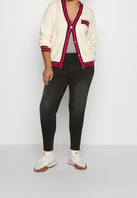 Gebreid crèmekleurig vest met rode en marineblauwe strepen op de kraag en manchetten, gecombineerd met zwarte slim-fit spijkerbroek en witte sportschoenen met bruine accenten.