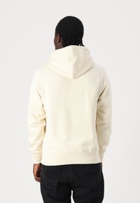 Felpa crema con cappuccio ampio, polsini e orlo a costine. Texture liscia, dettagli di cucitura minimi e vestibilità comoda. Pantaloni neri visibili.
